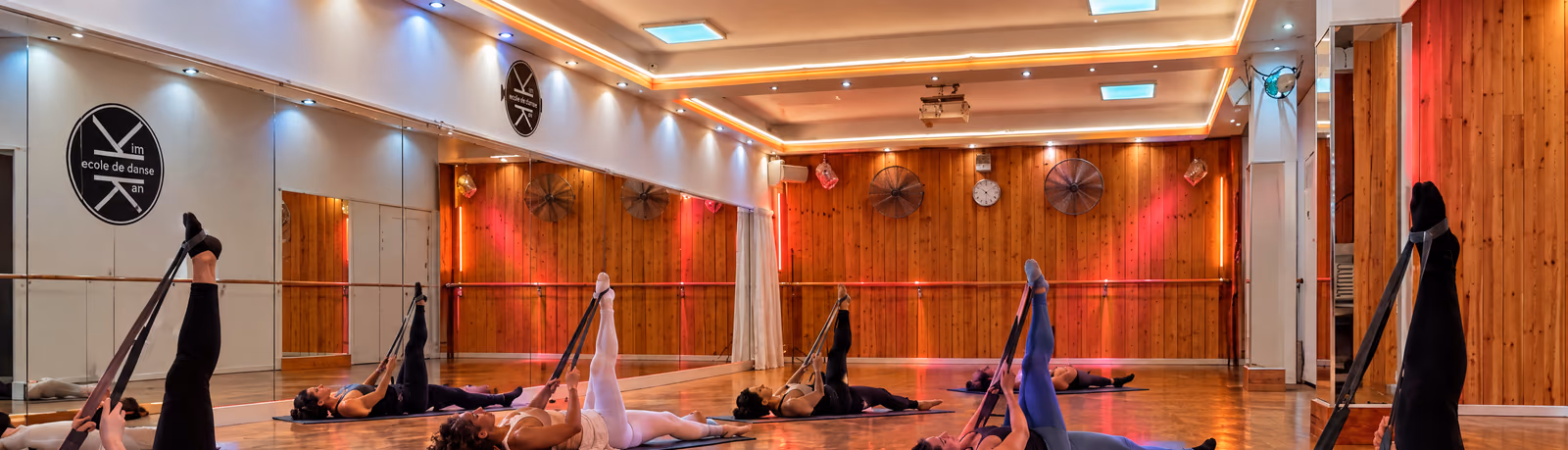 Barre au sol : des élèves allongés sur des tapis, une jambe tendue vers le plafond, dans la salle 2 de Gambetta, devant le lambris éclairé de rouge et le médaillon du logo.