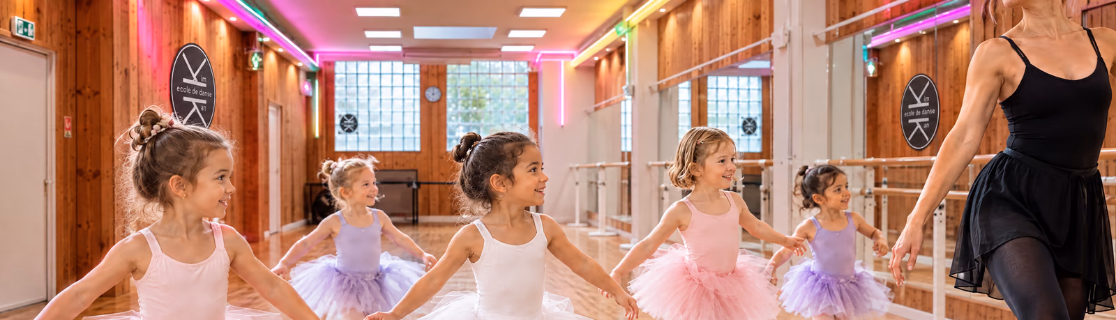 Éveil 4-5 ans : des fillettes en tutus roses, blancs et mauves avancent en file derrière leur professeure, dans la salle 1 de Gambetta, sous les rubans lumineux roses et verts.