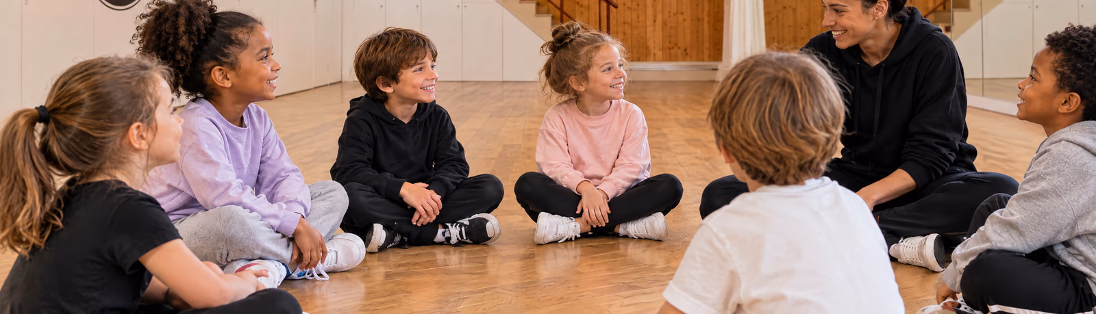 Initiation 6-7 ans : des enfants assis en cercle sur le parquet écoutent leur professeure, dans la salle 3 de Gambetta, devant l’escalier et le grand miroir.
