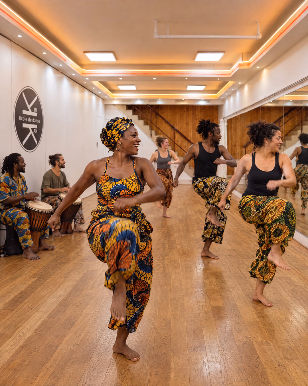 Danses africaines : des adultes en tenues wax lèvent les genoux, bras balancés, deux joueurs de djembé assis contre le mur, salle 3 de Gambetta.