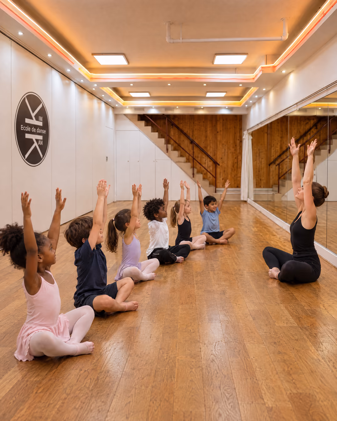 Initiation 6-7 ans : des enfants assis en rang sur le parquet, bras levés, face à leur professeure assise devant le miroir, dans la salle 3 de Gambetta.