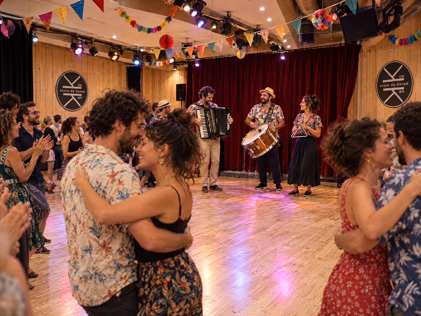 Bal de forró au Studio des Rigoles : un trio joue devant le rideau de velours rouge, accordéon, zabumba et triangle, sous les fanions ; deux couples dansent serrés au premier plan, on tape dans les mains autour d’eux.